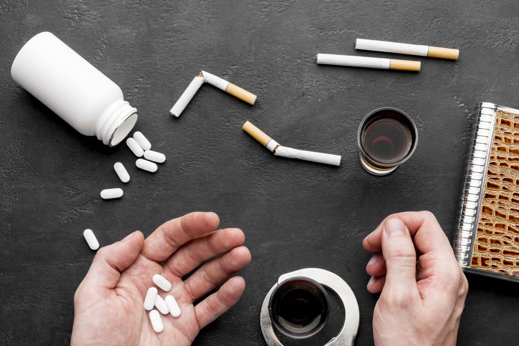 Hands near a spilling white pill bottle with several pills on a dark surface, surrounded by broken cigarettes, a glass of dark liquid, and a spiral notebook—conveying an addiction-related scene.