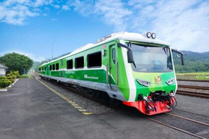 Green passenger train at a platform beside railway tracks on a sunny day with countryside in the background.
