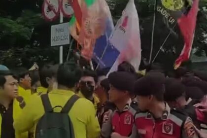 Crowd of people at an outdoor demonstration, holding colorful flags and banners with uniforms visible in the front.