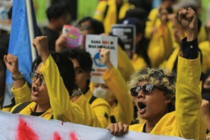 Protesters in yellow jackets raise fists, shouting slogans at a rally, holding signs and banners