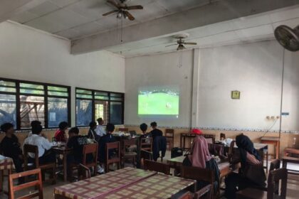 Group of students seated at tables in a classroom watching a soccer match projected on the wall.
