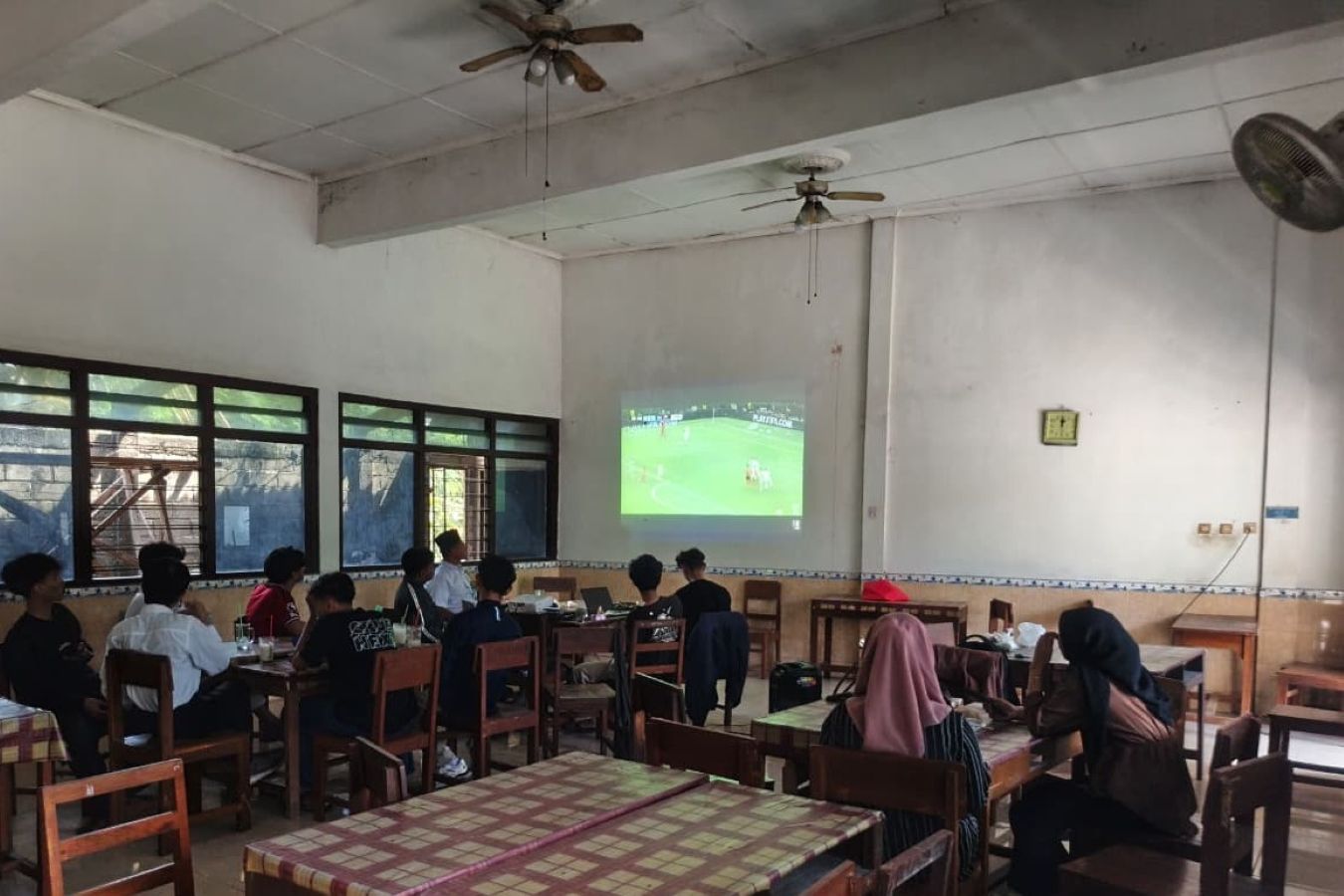 Group of students seated at tables in a classroom watching a soccer match projected on the wall.