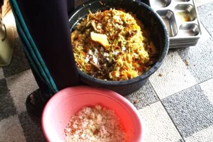 Person standing next to a large pot of mixed cooked food on a tiled floor; a pink bowl with salt sits nearby.