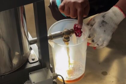 Person wearing gloves turns a red valve to release liquid into a translucent beaker on a lab bench area.