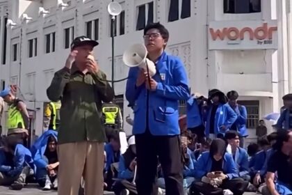 Man in blue jacket speaks into a megaphone while a man in olive green listens during a street protest, with a seated crowd in blue jackets behind them.