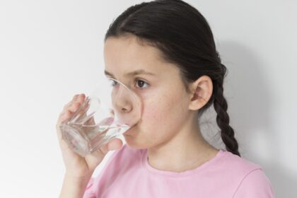Girl wearing a pink shirt drinking water from a clear glass.