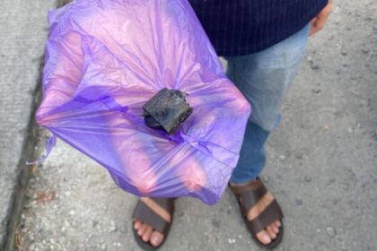 Person wearing sandals holds a crinkled purple plastic bag with a small black object on top, standing on a sidewalk.