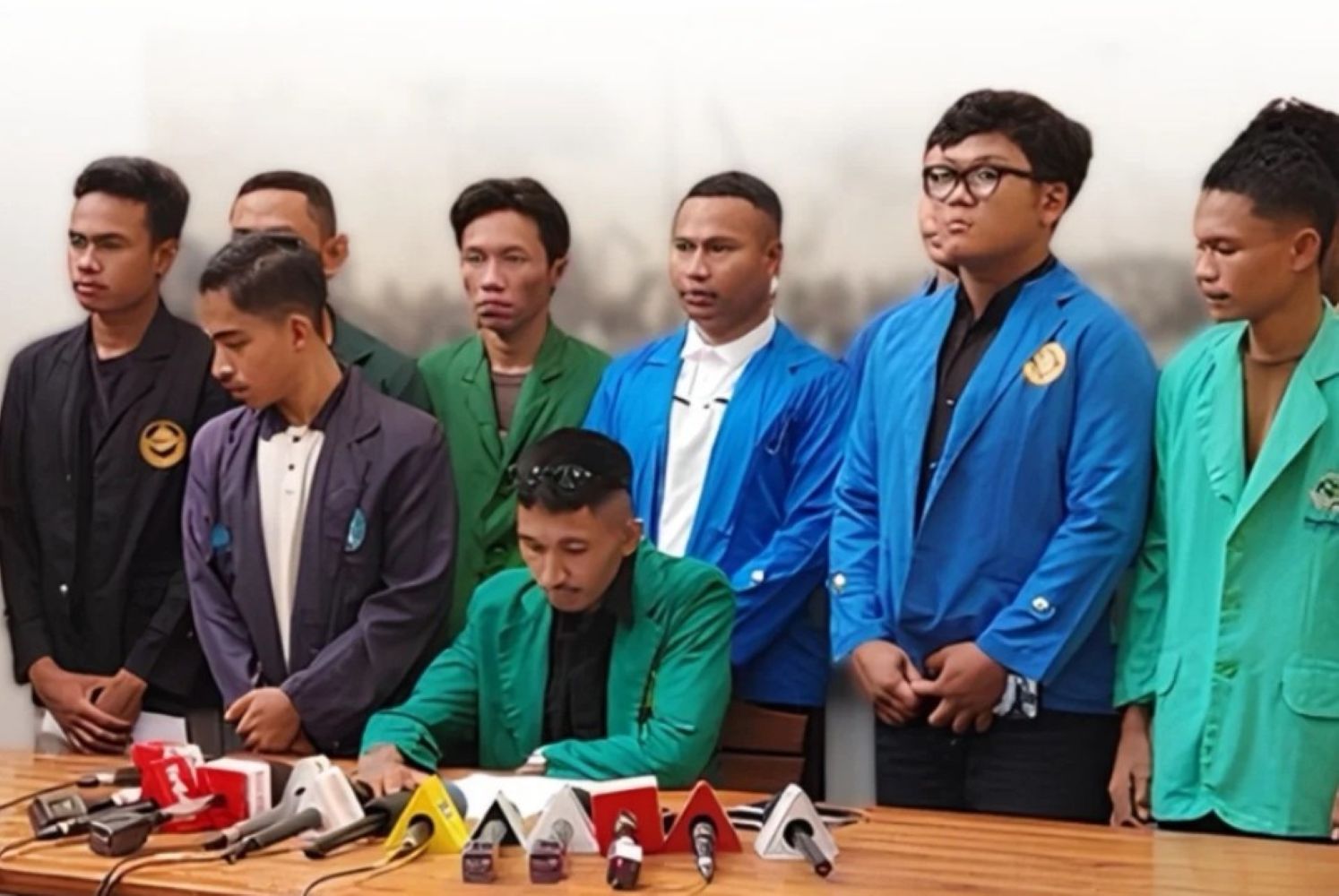 Man in a green blazer signs a document at a press conference while eight colleagues in colorful jackets stand behind him, microphones on the table.