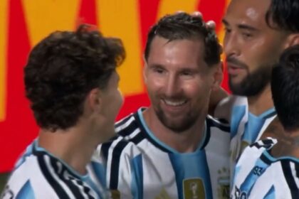 Two soccer players in Argentina national team jerseys celebrate together on the field, smiles visible amid a group celebration.
