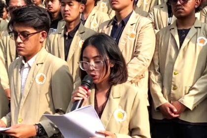Young woman with glasses speaks into a microphone while reading papers, surrounded by classmates in beige blazers at a school event.