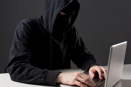 Person in a dark hoodie typing on a laptop, face obscured by the hood in a dim setting.