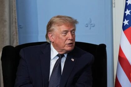 Older man in a dark suit and tie sits in a chair, with part of a US flag visible to the right.