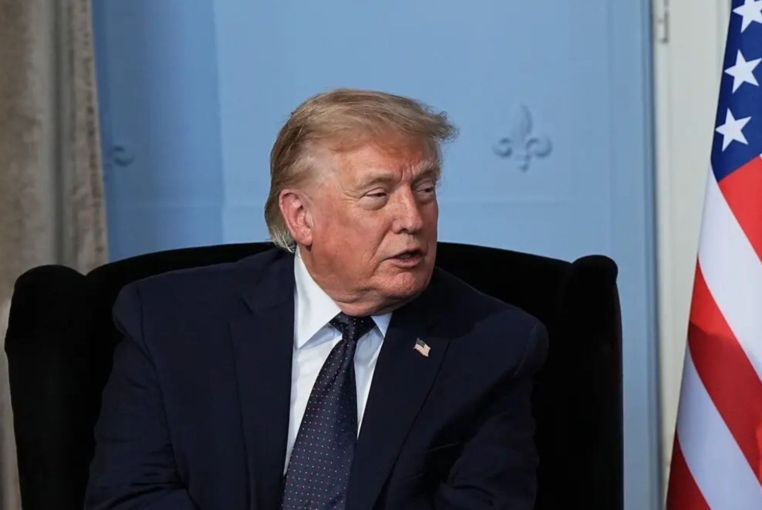 Older man in a dark suit and tie sits in a chair, with part of a US flag visible to the right.