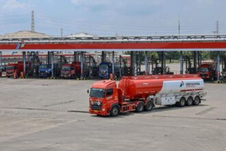 Red fuel tanker truck with a white trailer in the foreground at a Pertamina fuel depot; multiple loading bays and other trucks visible in the background.
