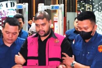 Man in a bright pink vest being escorted by two uniformed officers through a public area.
