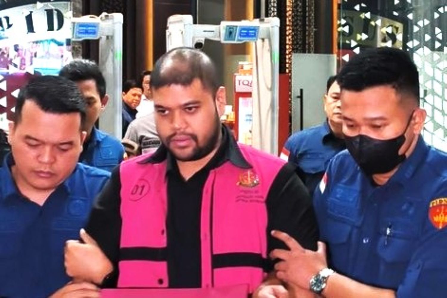 Man in a bright pink vest being escorted by two uniformed officers through a public area.
