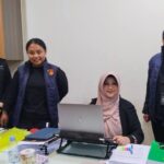 Four women in an office setting: one seated at a desk using a laptop, while three others stand beside her wearing blue padded vests with a crest.