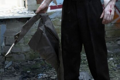 Person in black pants stands among rubble, gripping a torn dark metal sheet; blood on the right hand.
