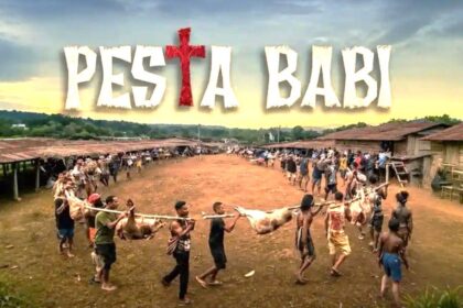 Festival banner reading 'Pesta Babi' above a village square as people carry a pig carcass on a pole in a long line of spectators and participants.
