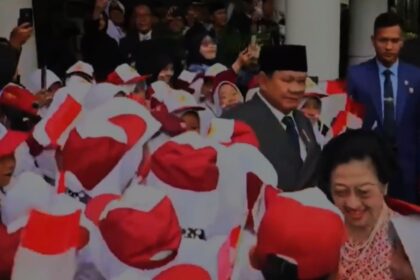 Crowd of people in red-and-white outfits waving Indonesian flags at a public ceremony; a smiling woman stands in the foreground.