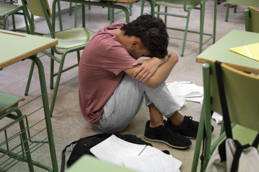 Student sitting on the classroom floor with head tucked into arms, surrounded by desks and scattered papers.