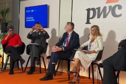 Panel of four professionals seated on stage in a discussion, PwC logo visible behind them and a blue screen reading 'Navigating Global Shifts' on the left.