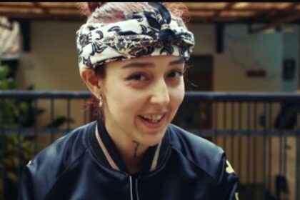 Young woman wearing a patterned headscarf and dark jacket smiles at the camera, holding a phone.