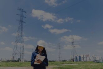 Woman in a white hard hat and yellow safety glasses writing in a notebook at a rural power-transmission site with tall lattice towers and a distant city skyline.