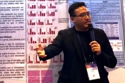 Male presenter in a black blazer speaks into a microphone in front of research posters with red bar charts behind him.