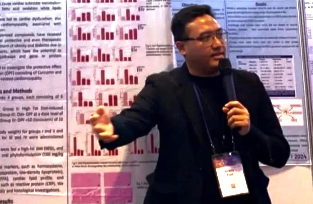 Male presenter in a black blazer speaks into a microphone in front of research posters with red bar charts behind him.
