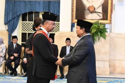Two men in suits wearing traditional black caps shake hands during a formal ceremony, with seated officials in the background and a portrait on the wall.