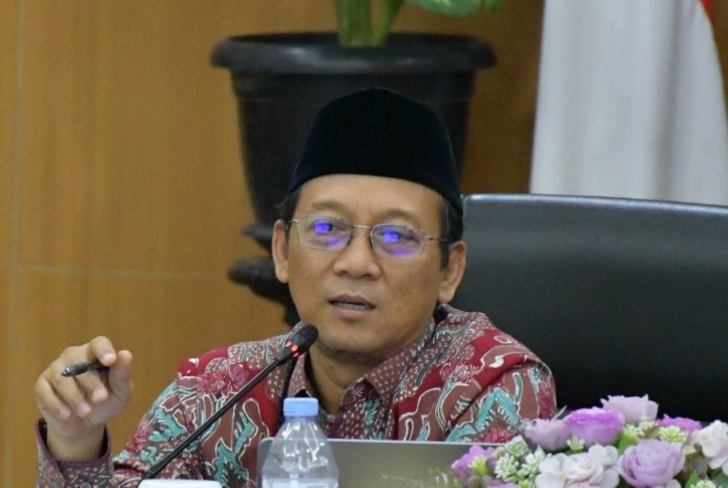 Asian man in a batik shirt and peci speaks at a panel with a microphone, laptop, water bottle, and flowers nearby.