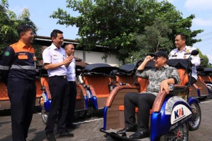 Man in a cap sits in a blue-and-orange pedicab while four uniformed men chat beside a row of orange rickshaws in a sunny parking lot.