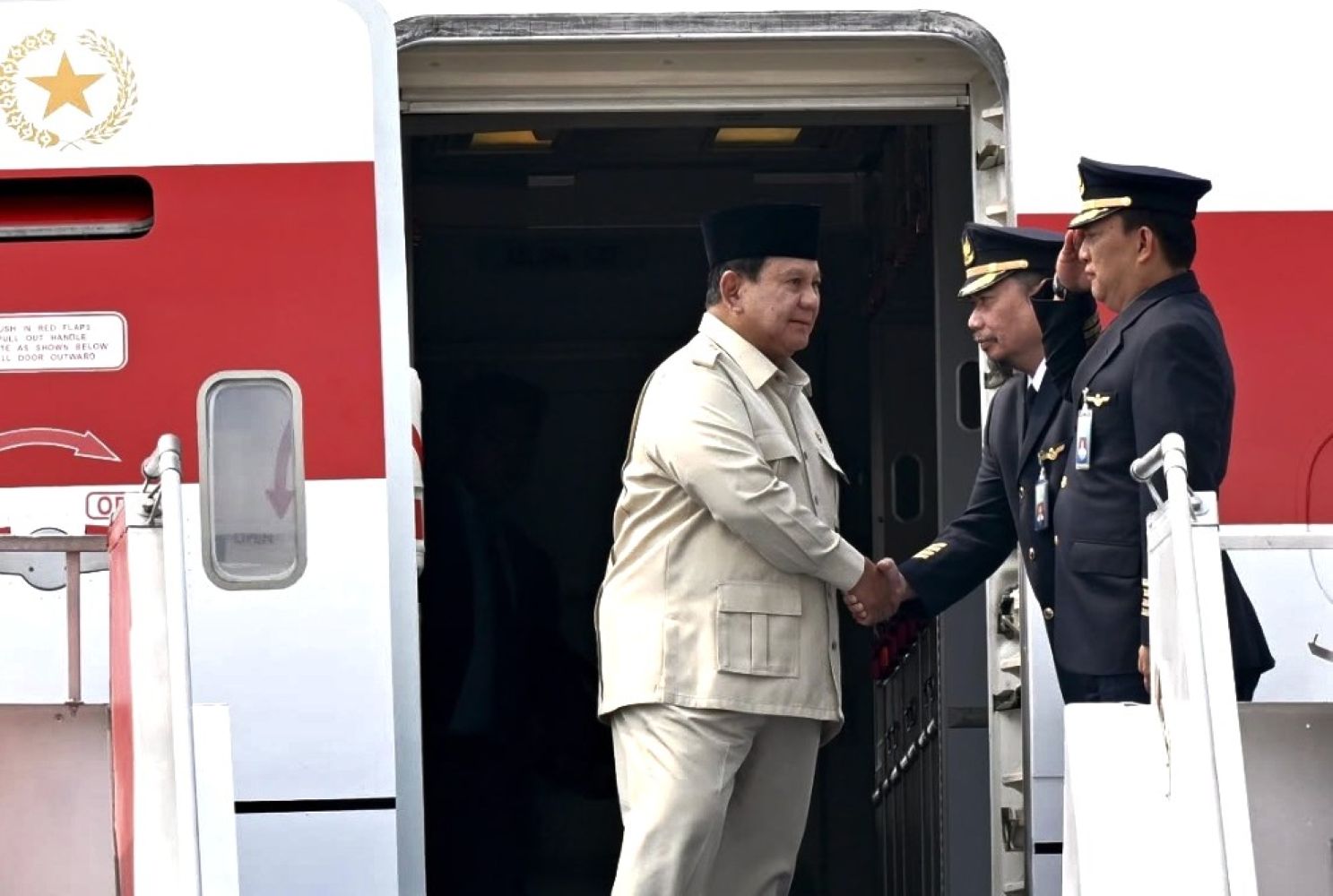 Two men shake hands at the open door of a red-and-white airplane, one in a beige suit and black cap, the other in a navy pilot uniform with gold badges.