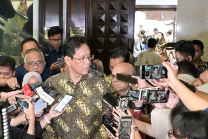 Man in a batik shirt speaks to reporters surrounded by microphones and smartphones in a crowded lobby corridor in front of a wooden door.