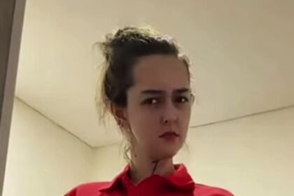 Close-up of a person with dark hair tied back, wearing a red shirt, looking to the side in an indoor setting.