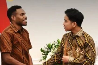 Two men in batik shirts shake hands in a formal setting, with flowers in the background.