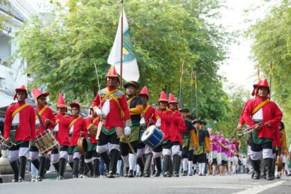 Parade of people in red uniforms marching with drums and a flag down a tree-lined street with spectators nearby.