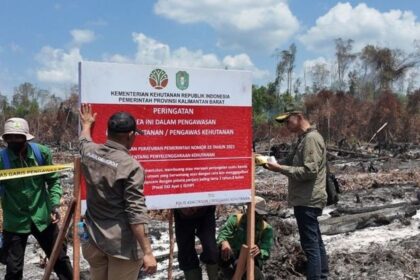 Officials in green uniforms inspecting a large red-and-white warning sign at a burned forest site, with caution tape and scorched ground in the background.