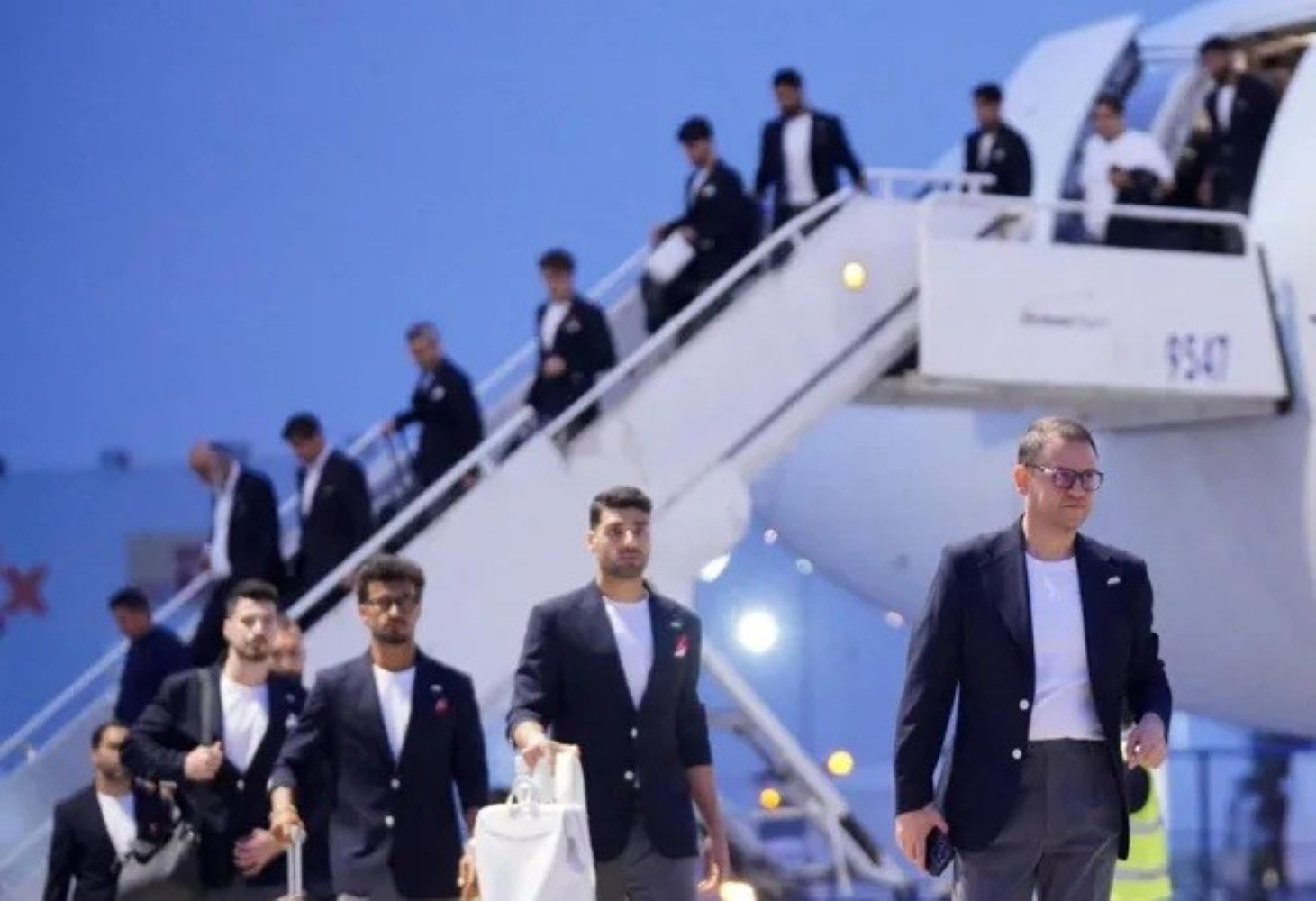 Group of men in dark blazers walking down stairs from a white airplane on the tarmac at the airport.