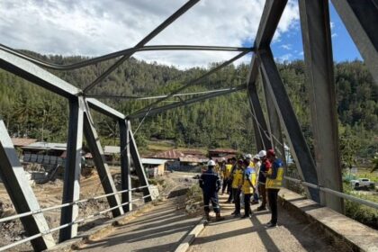 Group of construction workers in hard hats and yellow vests stand on a partially completed steel bridge, with a forested hillside in the background.