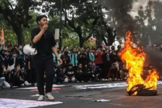 A young man in black speaks into a microphone as a pile of tires burns on a street protest, with a large crowd gathered behind him.
