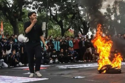A young man in black speaks into a microphone as a pile of tires burns on a street protest, with a large crowd gathered behind him.