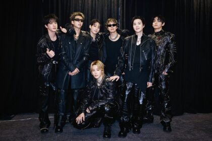 Seven men in black leather outfits posing together backstage against a dark curtain.