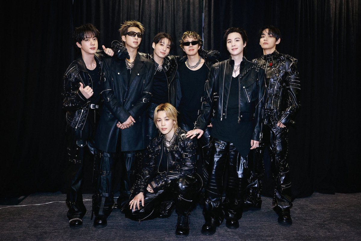 Seven men in black leather outfits posing together backstage against a dark curtain.