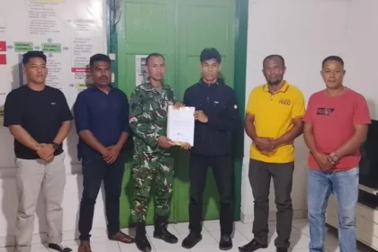 Six men pose indoors as a soldier in camouflage hands a document to a young man in black; others watch the handover.