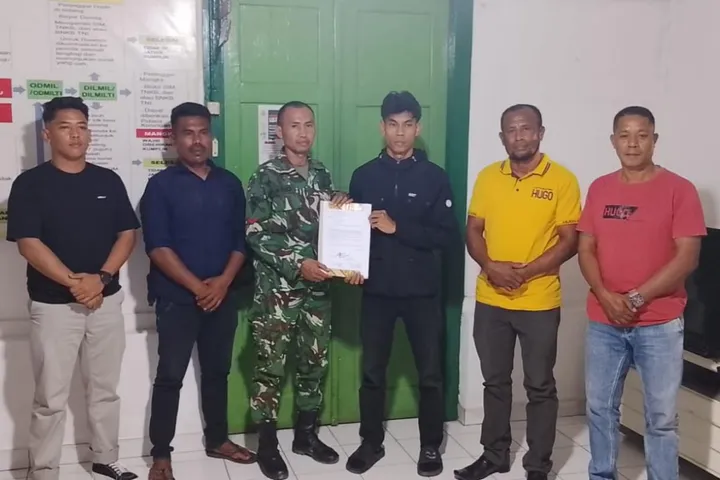 Six men pose indoors as a soldier in camouflage hands a document to a young man in black; others watch the handover.