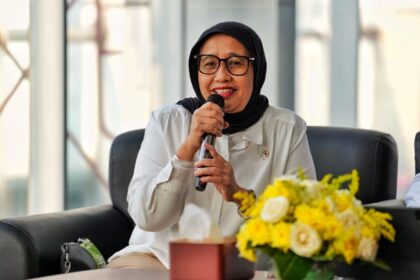 Woman wearing a black hijab and glasses speaks into a microphone during a panel, with yellow flowers on the table nearby.