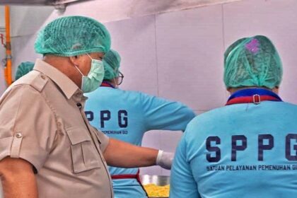 People in hairnets and face masks work at a food-preparation station, a supervisor in a tan uniform observes two staff wearing blue SPPG shirts beside a metal prep area.