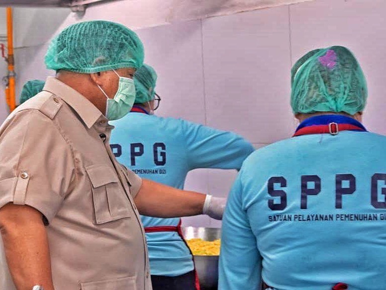 People in hairnets and face masks work at a food-preparation station, a supervisor in a tan uniform observes two staff wearing blue SPPG shirts beside a metal prep area.